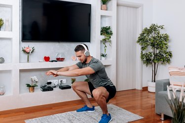 Adult performing kneel to squat exercise in living room while listening to music.