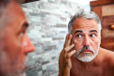 a person with grey hair and beard looking in the bathroom mirror and itching their face