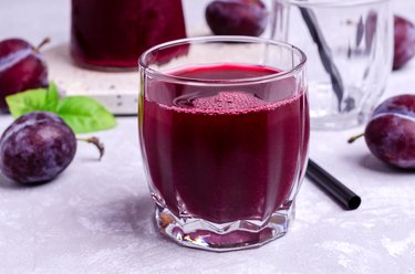 Red prune juice in a gladd next to fresh prunes on a table.