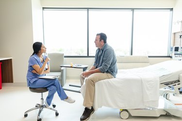 a doctor wearing blue scrubs and a brown ponytail sitting on a stool listening to a patient with short brown hair sitting on an exam table wearing a grey button-down shirt, khakis and sneakers