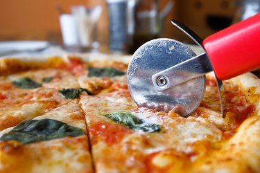 A close up photo of a margarita pizza with mozzarella, basil leaves and sauce on a wooden cutting board with a red pizza slicer rolling into a slice