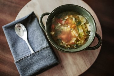 An overhead photo of a bowl of vegetable soup next to a napkin and spoon on a wooden table.