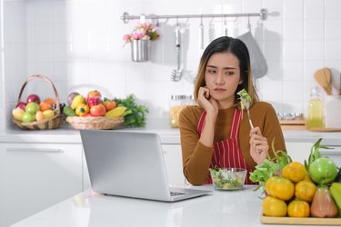 生气的年轻的亚洲女人吃沙拉。不满足的女人吃素食在使用笔记本电脑在厨房的桌子。raybet投注健康食品的概念。