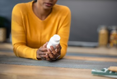 a person wearing a yellow longsleeve shirt holds a white pill bottle in a kitchen