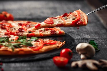 A slice of pizza on a spatula with smoked sausages, cheese, mushrooms, cherry tomatoes, bell peppers on a stone.