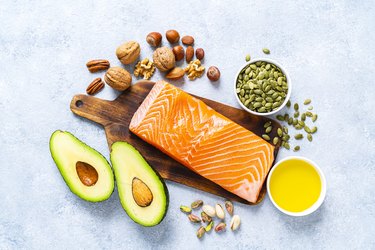 an overhead photo of foods high in healthy fats including salmon, avocado, olive oil, walnuts, pistachios, almonds and pumpkin seeds on a wooden cutting board on a white marble background