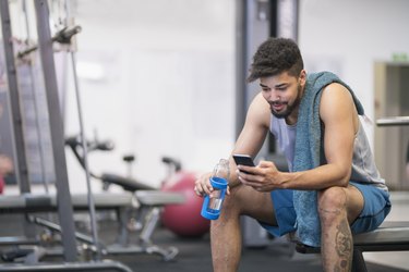 a person sits on a bench at the gym with a towel over their shoulder looking at their smartphone resting between sets