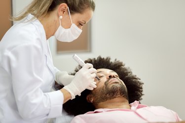 a person with a brown afro and beard getting a vampire facial from a dermatologist with a blonde ponytail wearing a surgical mask