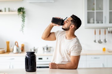 Young man drinking creatine from a shaker bottle in a kitchen