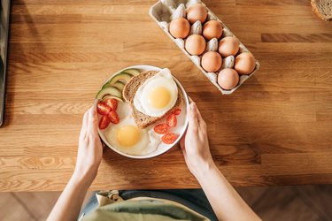 Person holding a plate of fried eggs, toast and vegetables next to a dozen eggs on a wooden table.