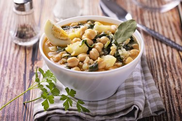 a white bowl of chickpea stew with spinach, hard boiled egg and a bay leaf on a checkered cloth napkin on top of a wooden table with a spoon and salt and pepper blurred out in the background