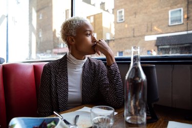 a person with short bleach blond curly hair wearing a grey sweater and white turtleneck and sitting in a restaurant thinking about the meal they just ate, to represent food noise