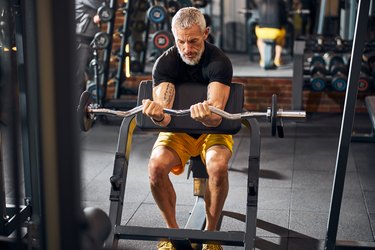 strong older man performing preacher curls with an EZ bar in a gym for bigger biceps