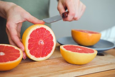 A girl or woman is holding a knife, cutting a red ripe grapefruit in half, on a cutting board, against the background of a wooden kitchen table. There is a manual juicer nearby. Making juice for breakfast. The concept of vegetarian, vegan and raw food.