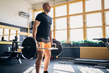 Person with short hair wearing orange shorts and a black t-shirt doing a deadlift with a hex bar at the gym