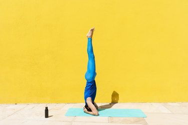 person in a blue tank top and blue leggings practicing Salamba Shirshasana while doing yoga by yellow wall