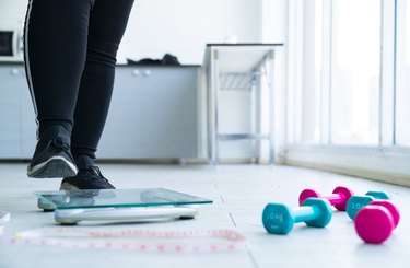 A close up of a person in black leggings and sneakers, stepping on a scale with weights in the background