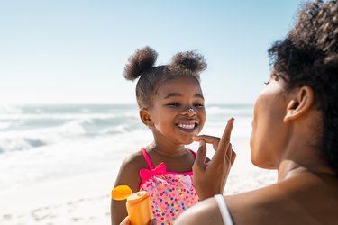 a parent applying sunscreen on a toddler in a pink bathing suit on the beach