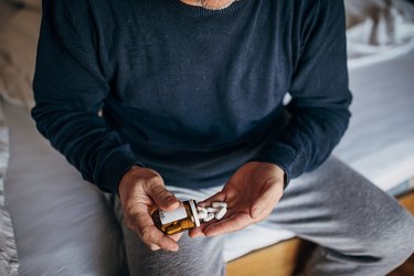 Man in grey sweatpants and sweatshirt sitting on bed pouring FertilAid for Men white pills into his hand, not ovaboost