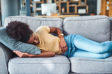 a person wearing light jeans and a yellow t-shirt lying down on a gray couch with stomach pain after eating spicy food