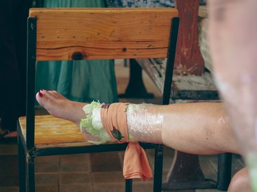 a photo of a person's leg propped on a wooden chair wearing anti-inflammatory cabbage leaves compressed with plastic wrap around their ankle