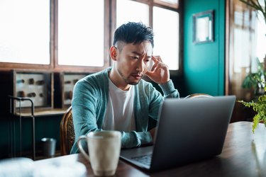 a person with short black hair sitting at a table and looking at a computer screen, looking concerned and stressed out