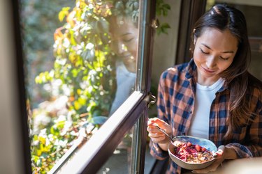 Woman smiling and eating bowl of berries and granola by sunny window wondering about the benefits of a 5-day fast bad breath during Ramadan
