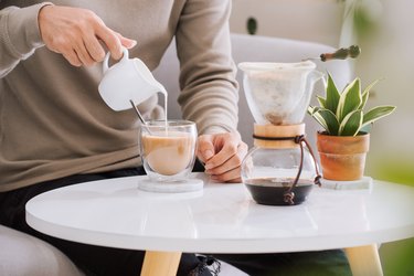 man's hand pouring sugary coffee creamer into a glass of espresso coffee in living room.