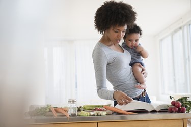 Mother reading cookbook with foods that increase milk supply while holding baby son