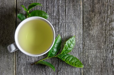 White cup of green tea with fresh tea leaves on wooden table, hot drink concept