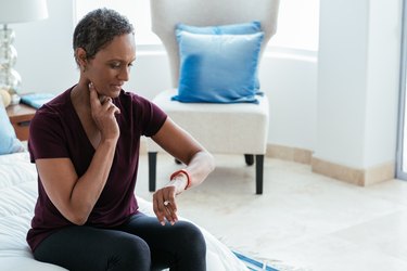 A person with short black, curly hair wearing a burgundy T-shirt and sitting on their bed while checking their pulse, to check their anxiety heart rate