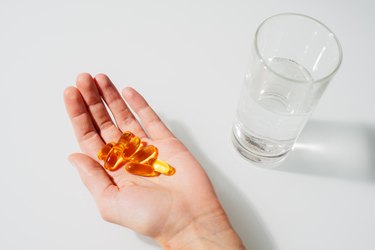Close-up of hand holding fish oil supplements and glass of water on white background