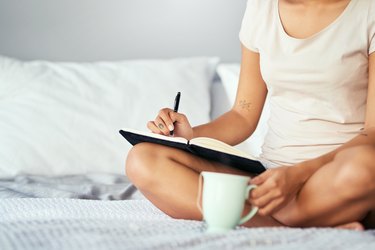 A woman sitting in bed drinking coffee and writing down her bathroom habits to track her gut health