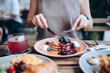 Close up of young woman sitting at dining table eating pancakes with blueberries and whipped cream in cafe