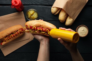 Person squeezes sauce on hot dog on wooden background, top view