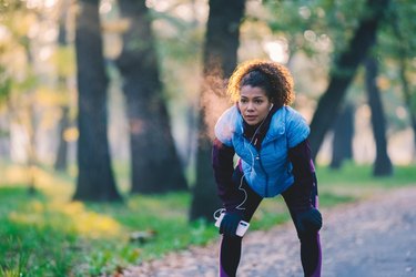 Woman in blue vest and black leggings on a forest path, working out on no sleep and listening to music on her phone through earbuds