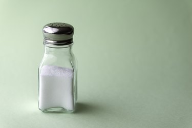 A glass shaker with a silver metal cap full of iodized salt, which can help with iodine consumption, on a light green background