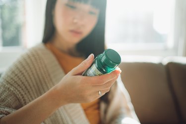 a person with shoulder-length dark hair and bangs wearing a beige sweater over an orange shirt looks closely at the label of a green supplement bottle in their hand to determine if there are any benefits for bloating and gas