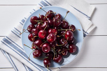 Top view of a plate of dark red cherries with a striped napkin on a white wooden table