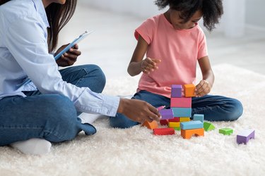 a cropped photo of a young kid and a child therapist playing with blocks on a white carpet