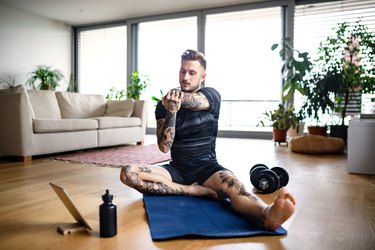 man with a flexibility fitness goal stretching in living room with a tablet, water bottle and dumbbells