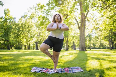 Young woman practicing tree pose (Vrksasana) with hands at heart center, doing yoga outdoors in park on a plaid blanket with a forest of green, leafy trees in the background
