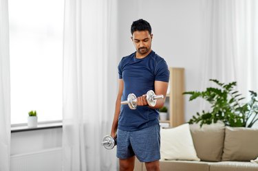Person wearing navy blue T-shirt and shorts doing biceps curl with dumbbell in living room.