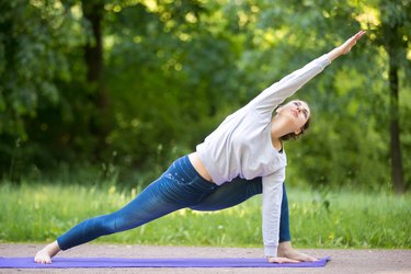 Young woman in blue leggings and a white sweater doing a side lunge stretch on a purple yoga mat outdoors
