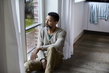 Pensive older adult sitting on the floor looking out of window