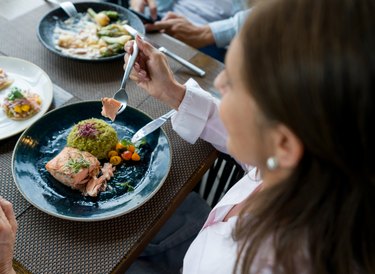 Top view of a happy, skinny mesomorph woman eating salmon, rice and veggies off of a blue plate at restaurant with other mesomorph guys