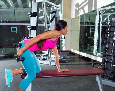 an athlete wearing a pink tank top and blue leggings does a dumbbell triceps kickback on a maroon bench at a gym