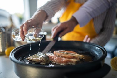 Senior woman pouring white wine into a pan of searing salmon, the best protein for longevity, while someone flips the fish with a metal spatula