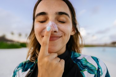 a person with long brown hair and blue fingernails standing on the beach and putting sunscreen on their nose