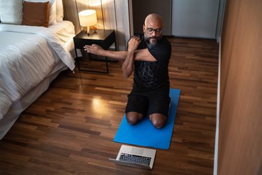 a person getting ready for a workout before bed on a blue yoga mat in a dimly lit bedroom in front of a laptop next to a bed and night table